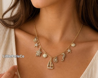 May include: Gold-toned necklace featuring various ocean-themed charms, including a sailboat, seahorse, starfish, and seashells. The necklace is worn by a person wearing a cream-colored top. The brand name "LittleParisCo" is visible on the left side.