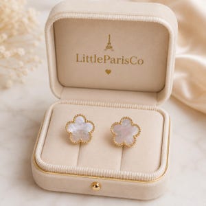 May include: A pair of gold-rimmed stud earrings shaped like five-petaled flowers with iridescent white mother-of-pearl centers. The earrings are displayed inside a beige velvet jewelry box featuring the text LittleParisCo and an Eiffel Tower icon.