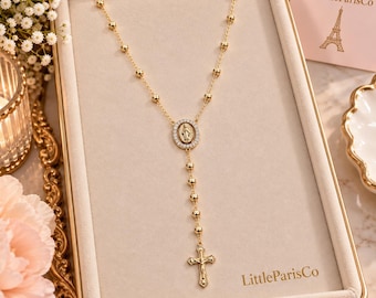 May include: A gold rosary necklace featuring a central oval pendant with a figure of the Virgin Mary surrounded by clear stones, a series of small gold beads along the chain, and a gold crucifix at the bottom, presented in a beige display box.