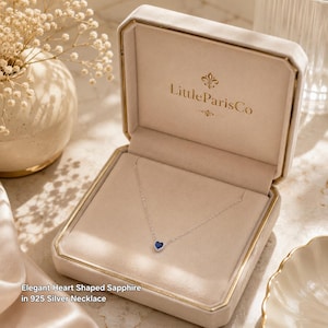 May include: A silver necklace with a heart-shaped blue sapphire pendant displayed inside a beige velvet-lined jewellery box. The interior lid features the gold text LittleParisCo. The text Elegant Heart Shaped Sapphire in 925 Silver Necklace is visible.