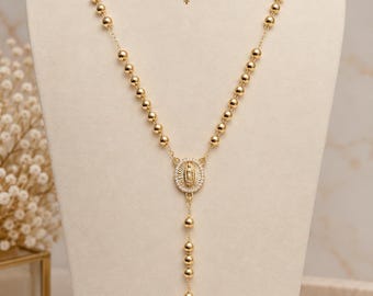 May include: A gold-toned rosary necklace featuring polished spherical beads, a central oval pendant with a figure surrounded by clear stones, and a crucifix at the bottom. The text LittleParisCo is printed above the necklace on the display.