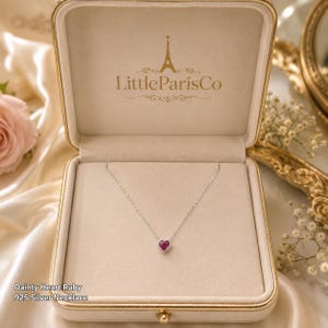 May include: A delicate silver necklace featuring a small, heart-shaped ruby pendant. The jewelry is presented inside a beige, square-shaped display box with the gold-colored text LittleParisCo and an Eiffel Tower icon printed on the inner lid.
