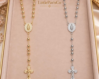 May include: Two rosary necklaces, one gold-toned and one silver-toned, each featuring a series of round beads, a central oval pendant depicting a figure, and a crucifix at the bottom. The text LittleParisCo is centered above the necklaces.