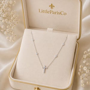May include: A silver necklace with a diamond cross pendant is displayed in a cream-colored jewelry box. The box is open, revealing the necklace with small, round diamonds. The box has gold trim and the text "LittleParisCo" is visible.