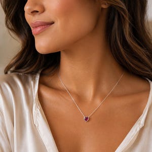 Puede incluir: Un collar de cadena plateada delicada con un pequeño colgante en forma de corazón que tiene una piedra preciosa de color rojo intenso en el centro. El colgante está enmarcado por un borde metálico fino y cuelga de una cadena fina.