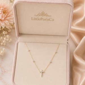 May include: A gold necklace with a cross pendant, displayed in a pink velvet jewelry box. The necklace features small, round gemstones and is presented against a cream-colored interior. The box is open, revealing the necklace.