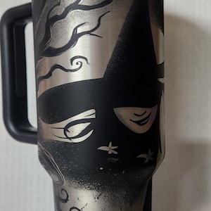 May include: A black stainless steel travel mug with a silver design of a face with a hat and a tree. The mug has a black handle and a black lid.