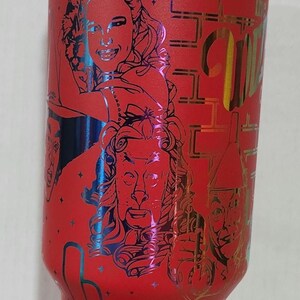 May include: Red stainless steel tumbler with a metallic iridescent finish. The tumbler features a graphic of characters from the Wizard of Oz, including Dorothy, the Cowardly Lion, the Tin Man, and the Scarecrow. The text "The Wizard of Oz" is printed on the tumbler.