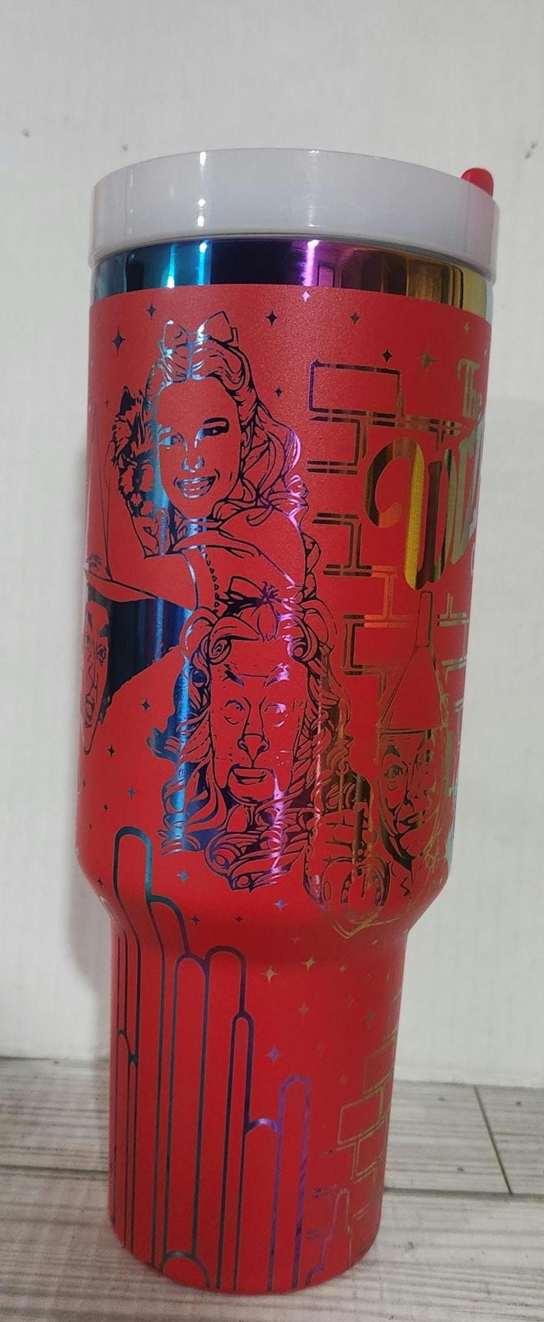 May include: Red stainless steel tumbler with a metallic iridescent finish. The tumbler features a graphic of characters from the Wizard of Oz, including Dorothy, the Cowardly Lion, the Tin Man, and the Scarecrow. The text "The Wizard of Oz" is printed on the tumbler.