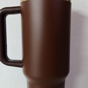 May include: A brown stainless steel travel mug with a gold band around the top and a black lid. The mug has a handle and is designed to keep drinks hot or cold.