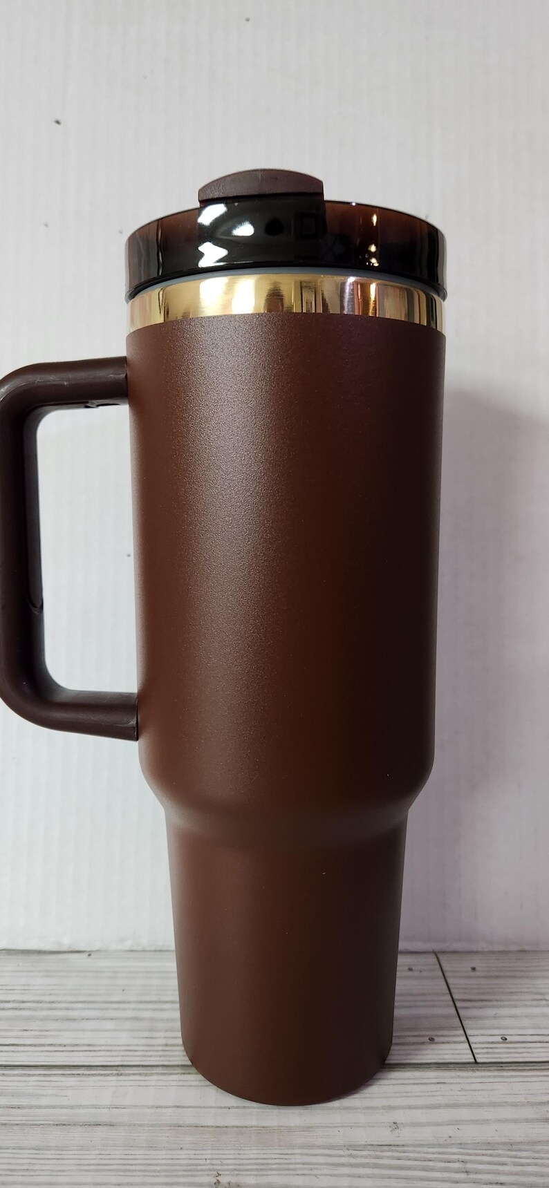 May include: A brown stainless steel travel mug with a gold band around the top and a black lid. The mug has a handle and is designed to keep drinks hot or cold.