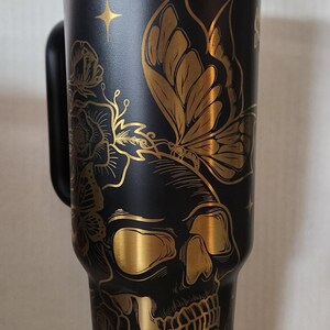 May include: A black and gold stainless steel tumbler with a skull and butterfly design. The tumbler has a lid and a handle.