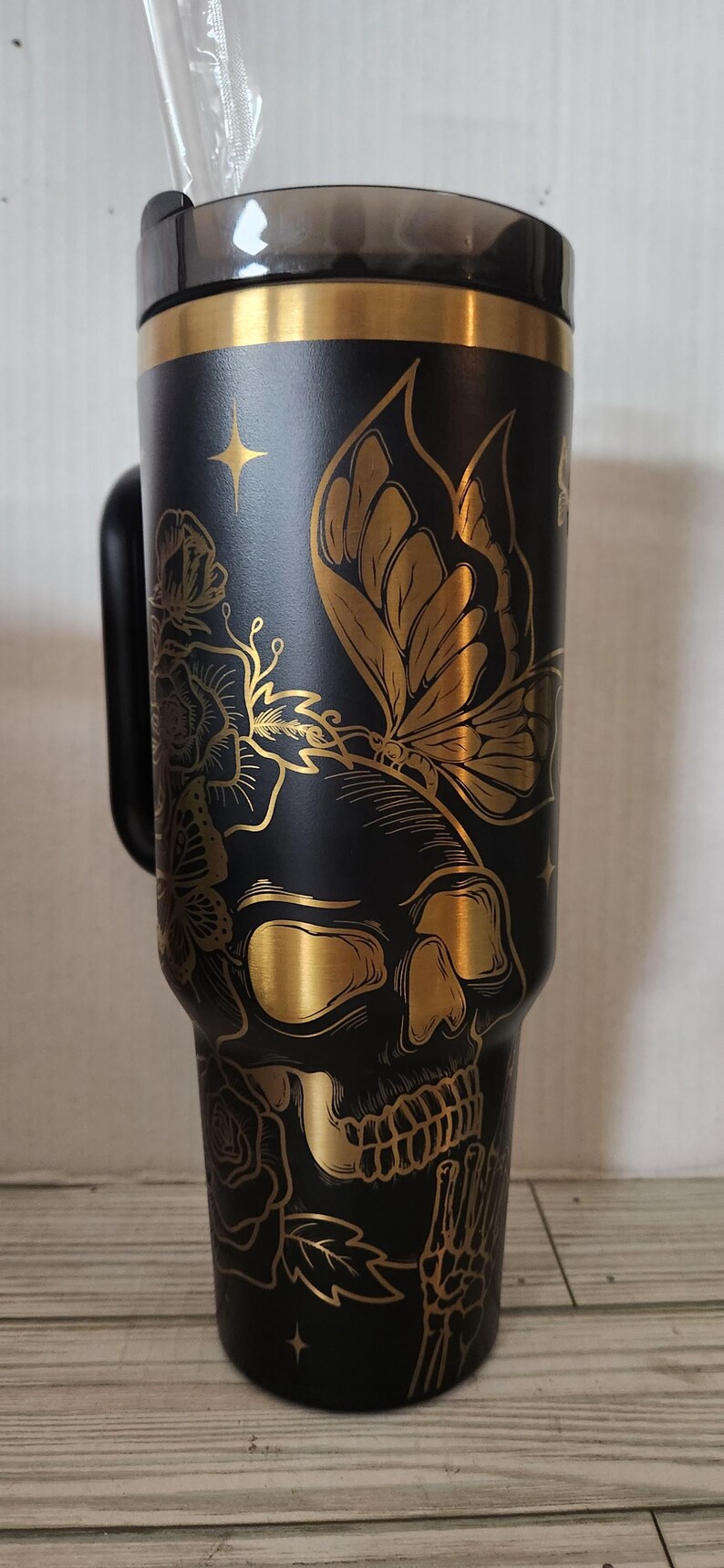 May include: A black and gold stainless steel tumbler with a skull and butterfly design. The tumbler has a lid and a handle.