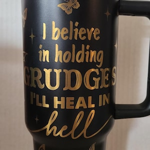 May include: Black travel mug with a gold inscription that reads "I believe in holding grudges I'll heal in hell". The mug has a black handle and a gold butterfly design.