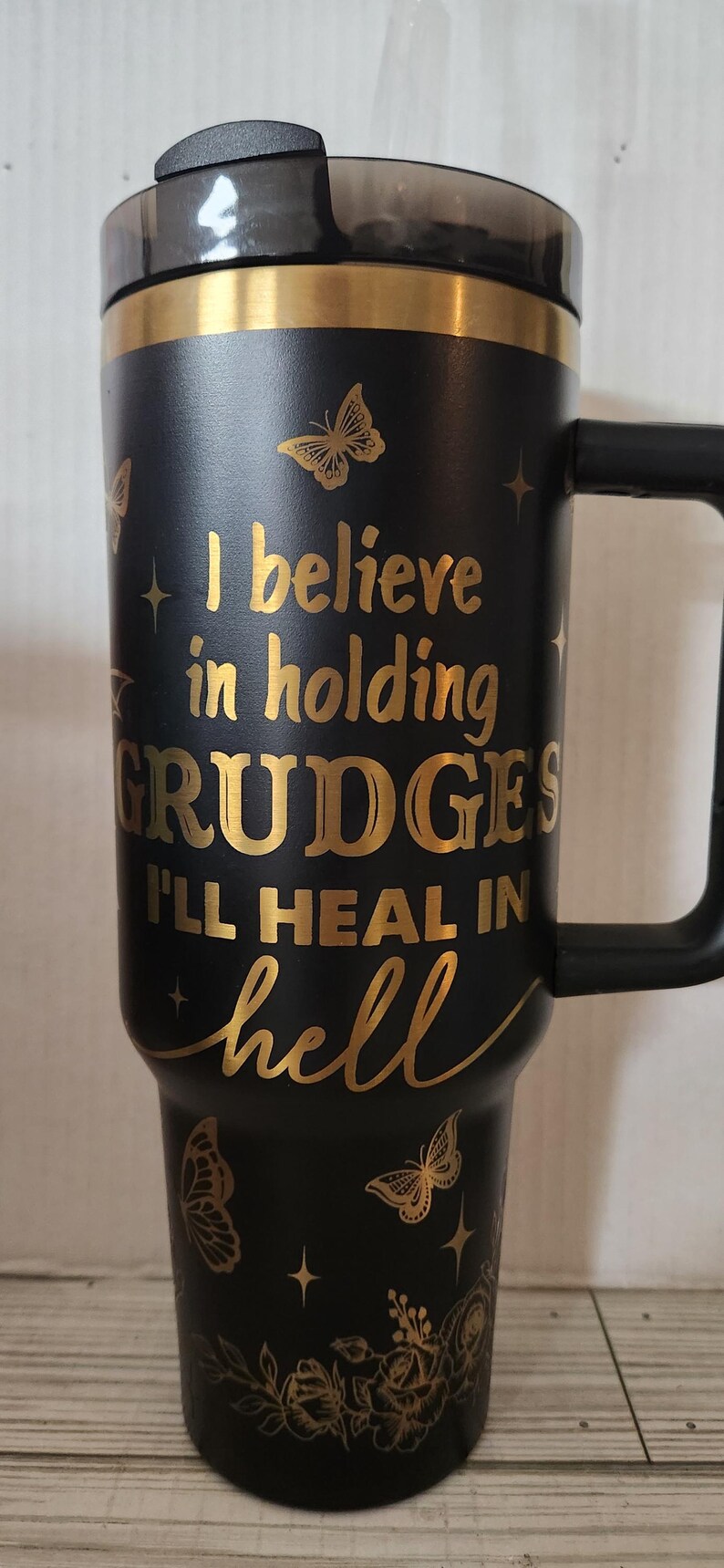May include: Black travel mug with a gold inscription that reads "I believe in holding grudges I'll heal in hell". The mug has a black handle and a gold butterfly design.