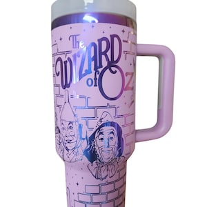 May include: Pink stainless steel travel mug with a handle. The mug features a graphic of the characters from the Wizard of Oz, including the Scarecrow, the Tin Man, and the Cowardly Lion. The text "The Wizard of Oz" is printed on the mug.