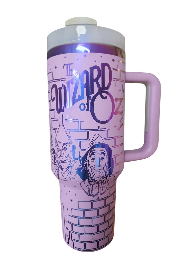 May include: Pink stainless steel travel mug with a handle. The mug features a graphic of the characters from the Wizard of Oz, including the Scarecrow, the Tin Man, and the Cowardly Lion. The text "The Wizard of Oz" is printed on the mug.