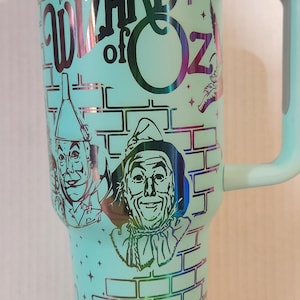 May include: A turquoise travel mug with a silver lid and handle. The mug features a graphic of the characters from the Wizard of Oz, including the Scarecrow, the Tin Man, and the Cowardly Lion. The text "The Wizard of Oz" is printed on the mug.
