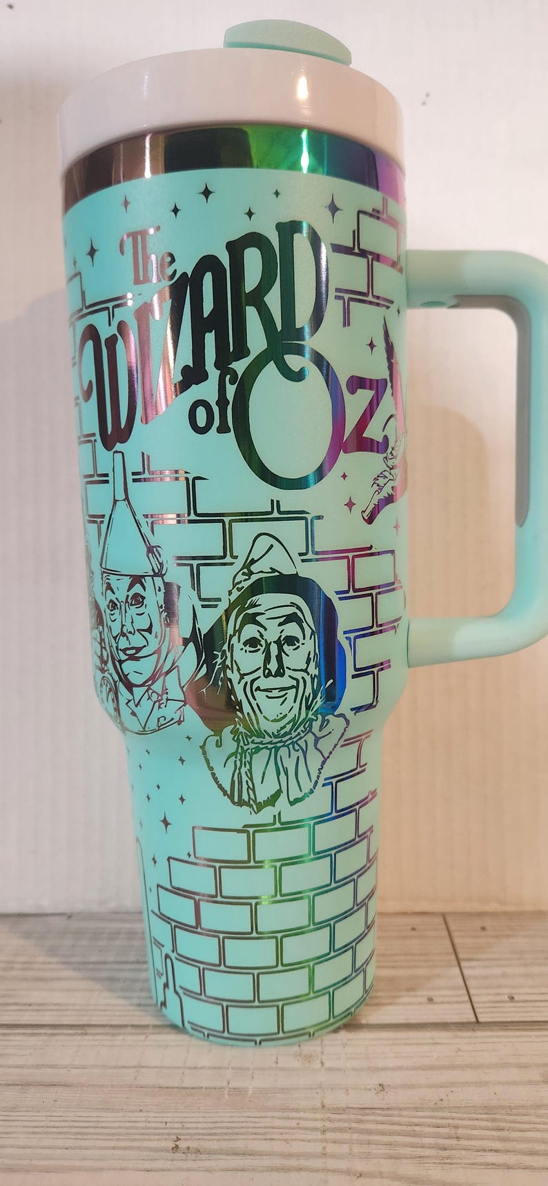 May include: A turquoise travel mug with a silver lid and handle. The mug features a graphic of the characters from the Wizard of Oz, including the Scarecrow, the Tin Man, and the Cowardly Lion. The text "The Wizard of Oz" is printed on the mug.