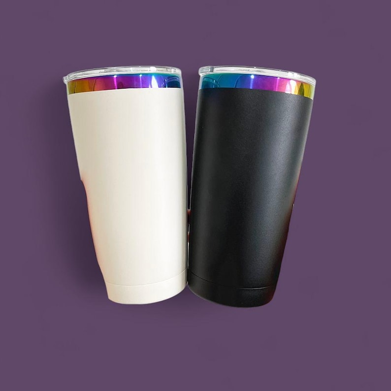 May include: Two insulated tumblers, one white and one black, with iridescent rainbow-colored rims. The tumblers are cylindrical and appear to be made of stainless steel. The background is a solid purple color.
