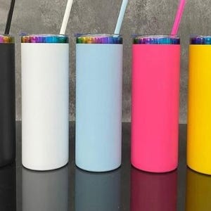 May include: Five colorful insulated tumblers with straws. The tumblers are black, white, light blue, pink, and yellow. Each tumbler has a rainbow-colored rim and a matching straw. The tumblers are cylindrical and appear to be made of metal.