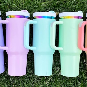 May include: Five pastel-colored insulated tumblers with handles are arranged on green grass. Each tumbler has a white lid with a rainbow-colored band and a matching handle. Colors include lavender, pink, teal, mint green, and peach.