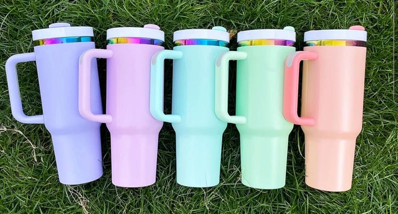 May include: Five pastel-colored insulated tumblers with handles are arranged on green grass. Each tumbler has a white lid with a rainbow-colored band and a matching handle. Colors include lavender, pink, teal, mint green, and peach.
