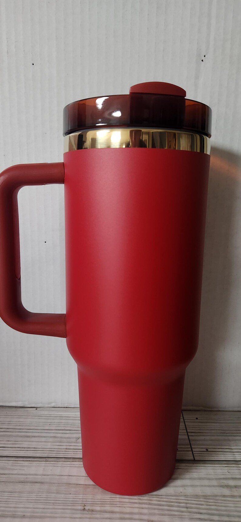 May include: A red travel mug with a gold rim and a brown lid. The mug has a handle and is made of stainless steel.