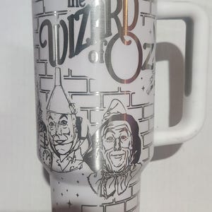 May include: White stainless steel travel mug with a black and white image of the characters from the Wizard of Oz. The mug has a black handle and the text "The Wizard of Oz" is printed on the front.