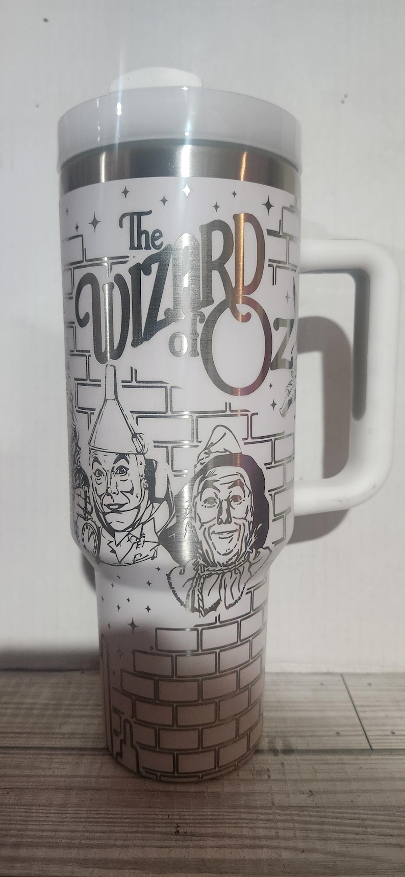 May include: White stainless steel travel mug with a black and white image of the characters from the Wizard of Oz. The mug has a black handle and the text "The Wizard of Oz" is printed on the front.