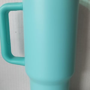 May include: A turquoise travel mug with a gold band around the middle and a green lid. The mug has a handle and is made of stainless steel.