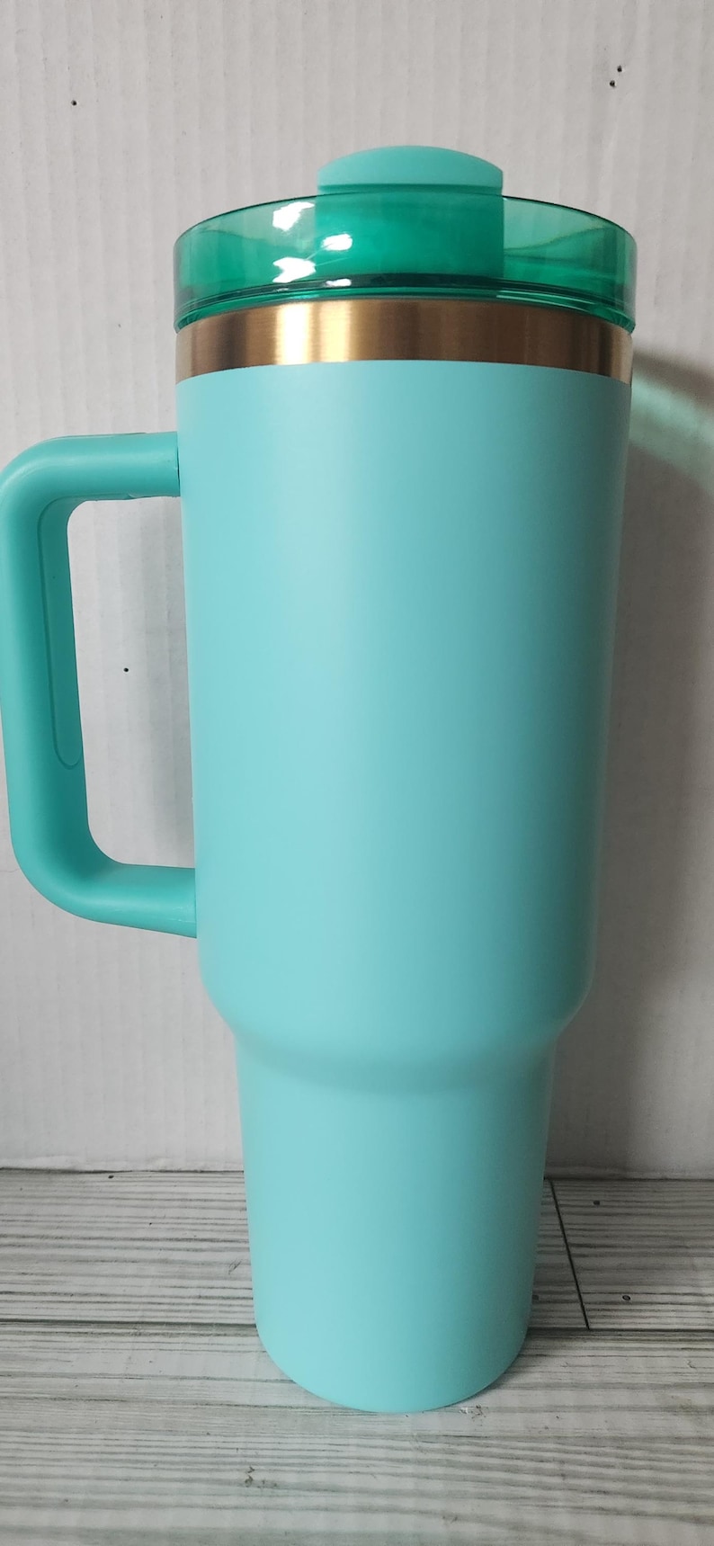 May include: A turquoise travel mug with a gold band around the middle and a green lid. The mug has a handle and is made of stainless steel.