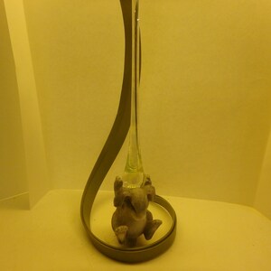 May include: A decorative glass vase with a light green tint, featuring a gray metal loop and a small, detailed figurine of an animal. The vase has a slender, elongated shape, and the figurine is positioned near the base.