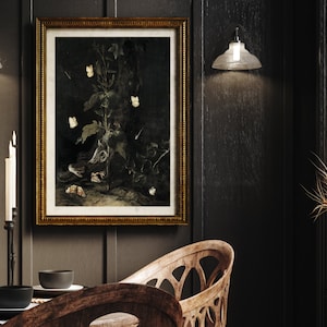 May include: A framed black and white still life print of a plant with white flowers and butterflies. The print is in a gold frame and is hanging on a dark wall. There are candles and a table setting in the foreground.