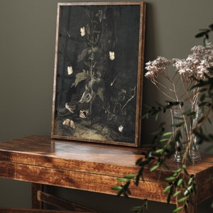 May include: A framed print of a dark, moody still life featuring a plant with white butterflies and a brown, textured surface. The print is in a wooden frame and is hanging on a dark green wall.
