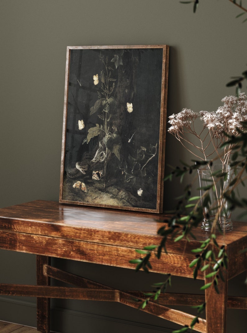 May include: A framed print of a dark, moody still life featuring a plant with white butterflies and a brown, textured surface. The print is in a wooden frame and is hanging on a dark green wall.