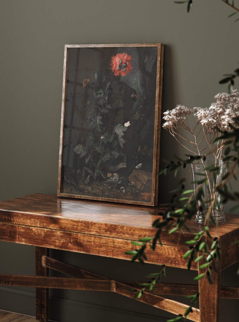 May include: A framed print of a still life featuring a red poppy flower, green leaves, and insects. The print is in a wooden frame and is hanging on a dark green wall.
