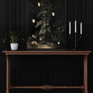 May include: A dark, framed painting of a still life featuring a snake, a skull, and a plant with white butterflies. The painting is hanging on a black wall above a wooden console table with a potted plant.