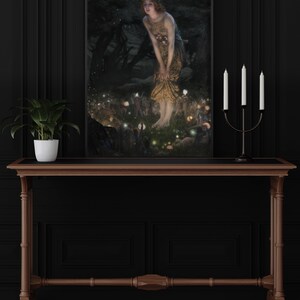 Op de afbeelding: Een ingelijst schilderij van een vrouw in een bosrijke omgeving. De vrouw draagt een lange, vloeiende jurk en is omringd door kleine, gloeiende lichtjes. Het schilderij heeft een donkere achtergrond.
