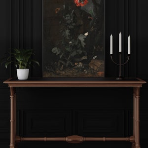 May include: A framed painting depicting a red poppy flower with green leaves against a brown background. The painting is hung on a black wall above a wooden table with a plant in a white pot and a black candle holder with three white candles.