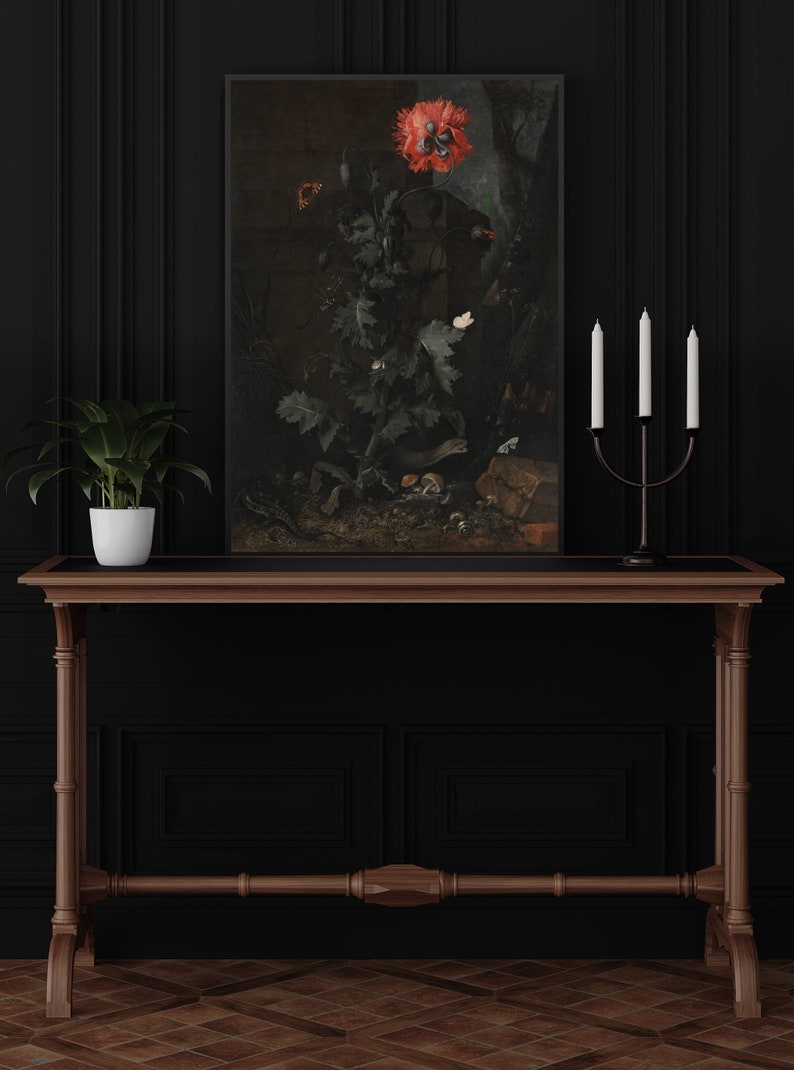 May include: A framed painting depicting a red poppy flower with green leaves against a brown background. The painting is hung on a black wall above a wooden table with a plant in a white pot and a black candle holder with three white candles.
