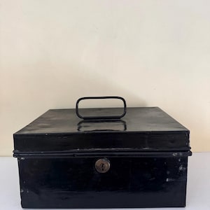 Puede incluir: Una caja registradora de metal negro con forma rectangular. Cuenta con un asa en la parte superior y una cerradura en la parte delantera. La caja presenta un aspecto ligeramente desgastado, lo que sugiere edad y uso. El dise&ntilde;o es simple y funcional.