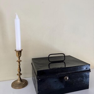Puede incluir: Un candelabro de lat&oacute;n vintage con una vela blanca junto a una caja met&aacute;lica negra con asa y cerradura. Los objetos est&aacute;n sobre una superficie blanca contra una pared crema. Estilo cl&aacute;sico.