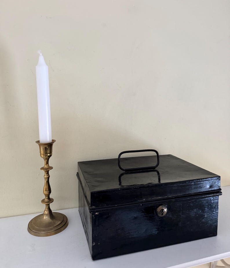 Puede incluir: Un candelabro de lat&oacute;n vintage con una vela blanca junto a una caja met&aacute;lica negra con asa y cerradura. Los objetos est&aacute;n sobre una superficie blanca contra una pared crema. Estilo cl&aacute;sico.