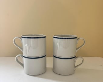 May include: Four white ceramic mugs stacked in pairs. Each mug has a dark blue band around the top and another around the middle. The mugs have curved handles and are set against a neutral background.