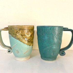 May include: Two ceramic mugs with unique designs. One mug is light blue with brown accents and the other is a deep teal blue.