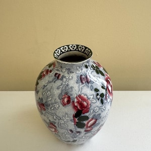 May include: A round ceramic vase with a floral pattern. The vase features a light blue background with red roses and green leaves. The rim is dark green with a white decorative design.
