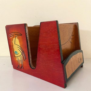 May include: A wooden napkin holder with a red, orange, and yellow painted design of a bird. The holder has a black trim and a signature on the bottom.