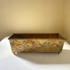 May include: A rectangular brass planter featuring an embossed floral vine pattern along the exterior sides. The metallic container has a warm, aged patina finish and is supported by four small, rounded feet at the base corners.