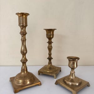 May include: Three antique brass candlesticks of varying heights. Each candlestick has a square base and a detailed, ornate design. The tallest candlestick is approximately 10 inches tall, while the shortest is about 4 inches.